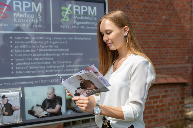 Eine Frau steht vor einem Infoschild von RPM Medical & Kosmetik® und schaut in eine Broschüre